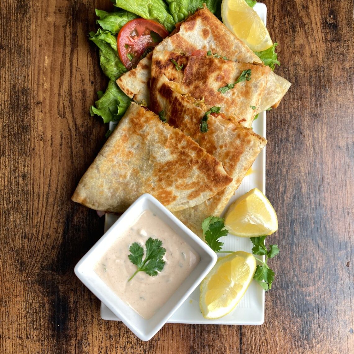 Golden-brown crispy chicken quesadilla triangles served on a white platter with a creamy dipping sauce, fresh lemon wedges, and a side of lettuce and tomato.