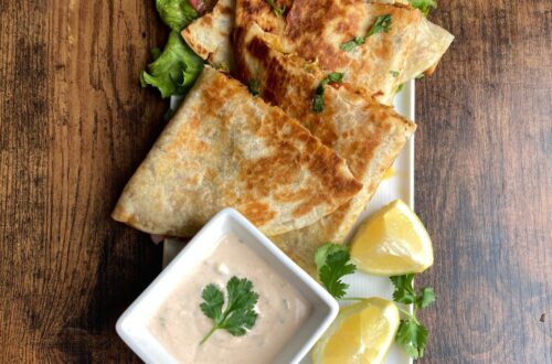 Golden-brown crispy chicken quesadilla triangles served on a white platter with a creamy dipping sauce, fresh lemon wedges, and a side of lettuce and tomato.