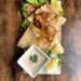 Golden-brown crispy chicken quesadilla triangles served on a white platter with a creamy dipping sauce, fresh lemon wedges, and a side of lettuce and tomato.